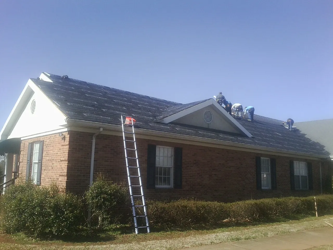 Crew tearing shingles off a brick ranch roof with a ladder set — Metal Roof Repair work by our crew in Huffman