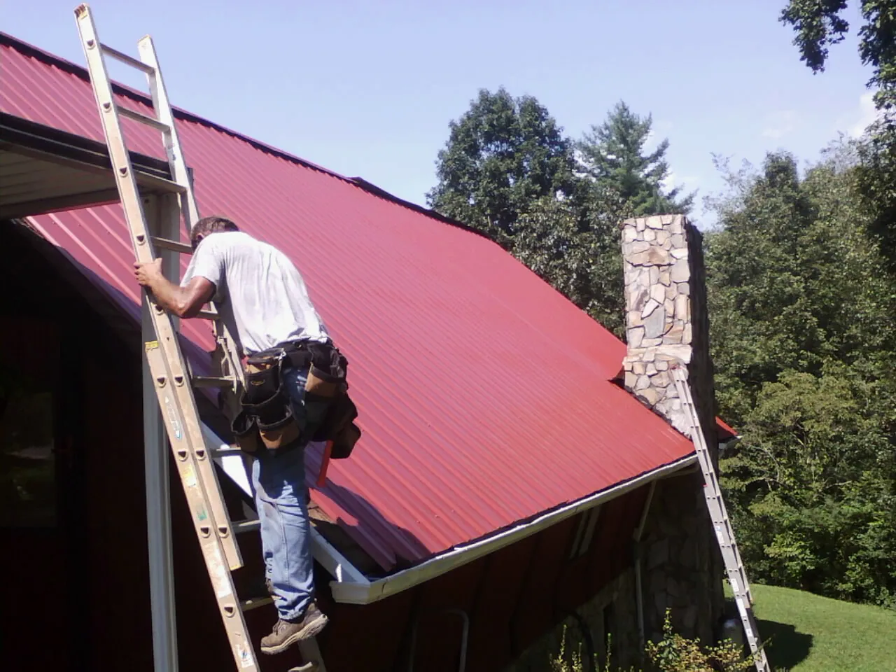 Roofer on a ladder beside a red metal roof and stone chimney — Soffit & Fascia Repair by Roofing Brothers in Huffman